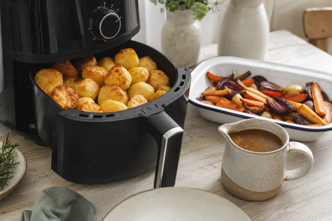 Air fryer in a bright kitchen next to potatoes, chicken thighs, roasted vegetables and a sponge cake