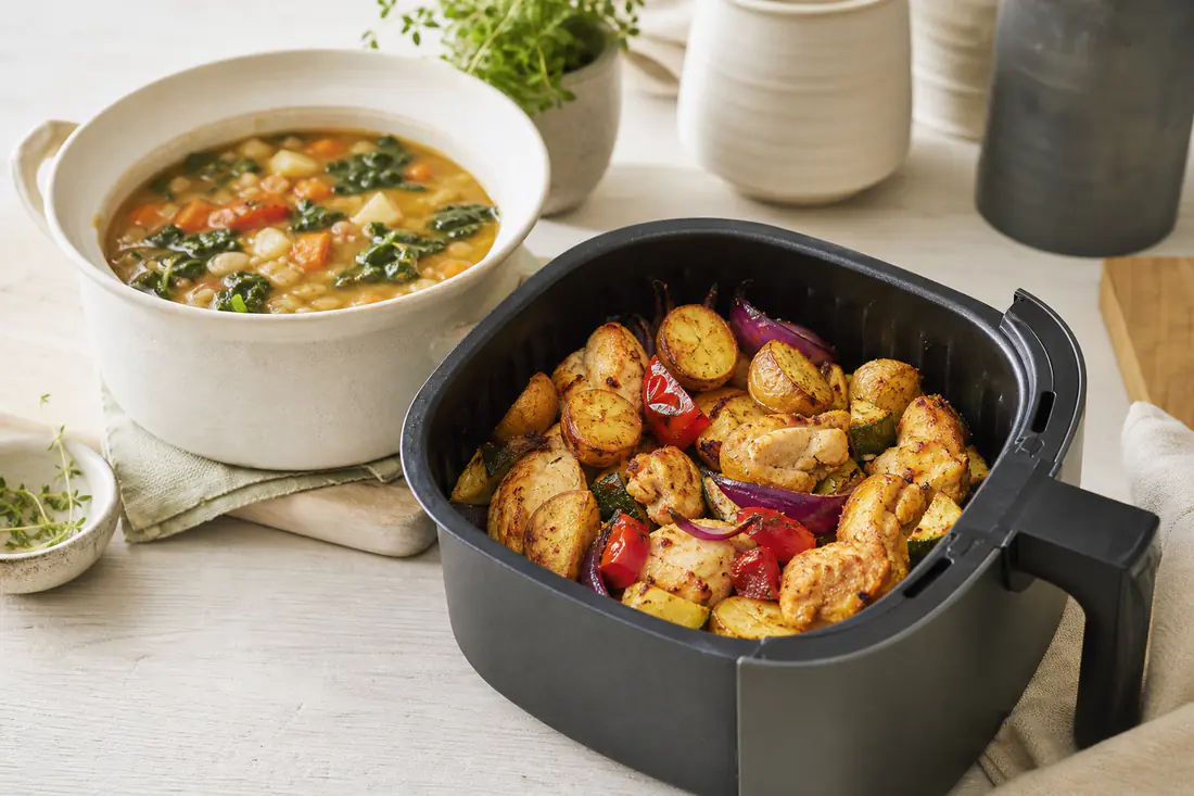 Crispy potatoes, chicken pieces and vegetables in an air fryer basket next to a pot of soup