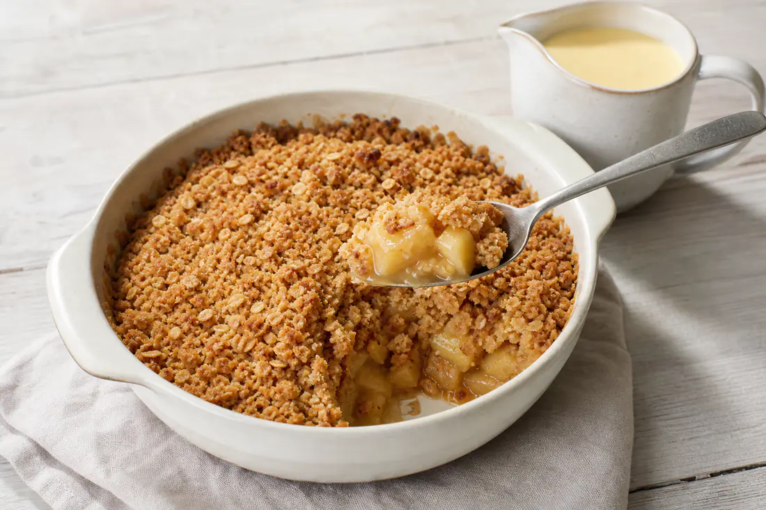 A golden apple crumble in a small ovenproof dish with a spoonful lifted out beside a jug of custard