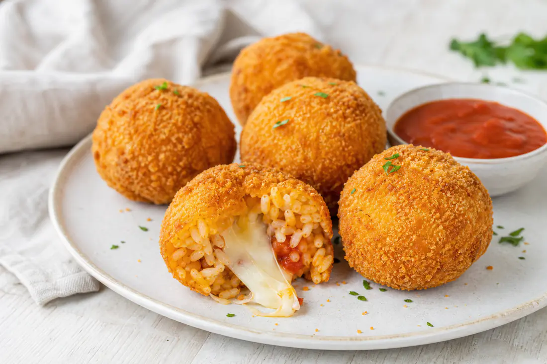 Golden breadcrumbed arancini rice balls, one split open to show melted mozzarella, on a plate with tomato sauce