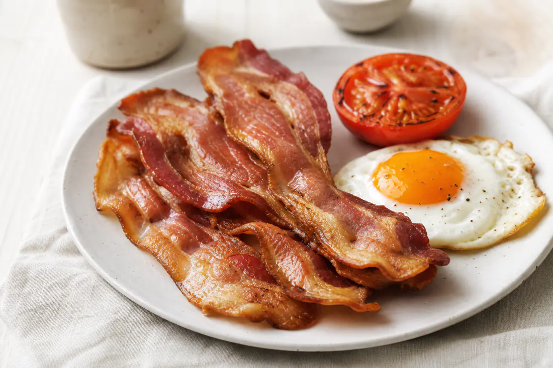 Golden crispy streaky bacon rashers piled on a white plate next to a fried egg and grilled tomato
