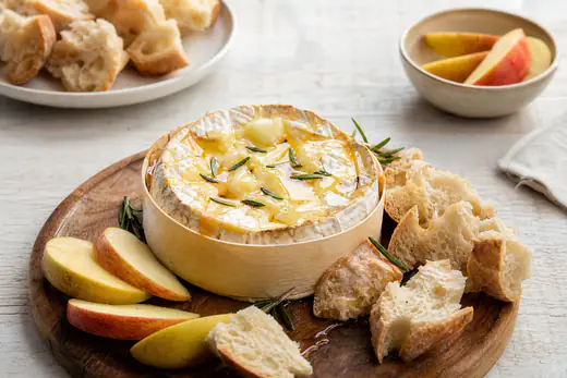 Baked camembert with a cut-open top, molten centre, honey and rosemary on a board with torn bread and apple slices