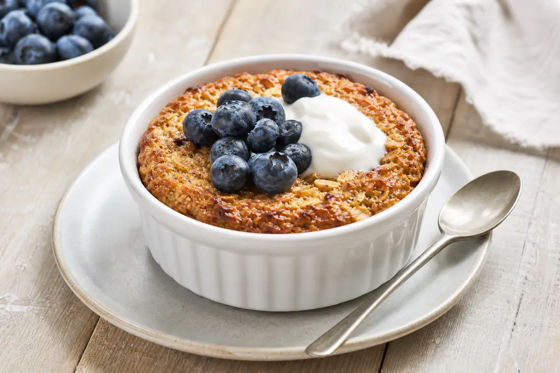 A white ramekin of golden baked oats with blueberries and a spoonful of yoghurt on a wooden table