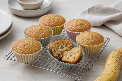 Six golden banana muffins in silicone cases on a wire rack, one split open to show the moist crumb, with a ripe banana beside them
