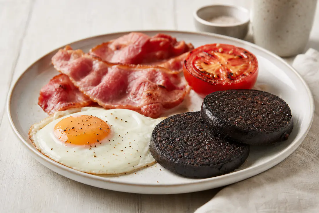 Crisp slices of black pudding on a plate with a fried egg, bacon and grilled tomato for a full English breakfast