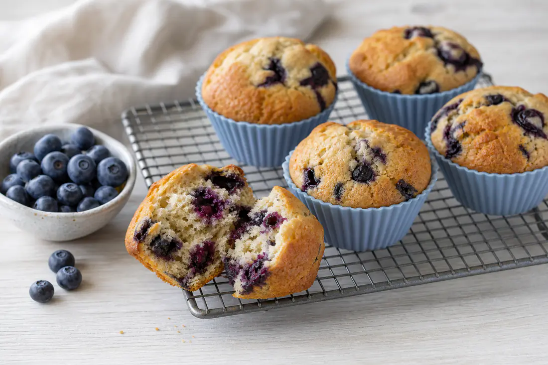 Golden blueberry muffins in silicone cases on a wire rack, one broken open to show purple berries inside, with a small bowl of fresh blueberries