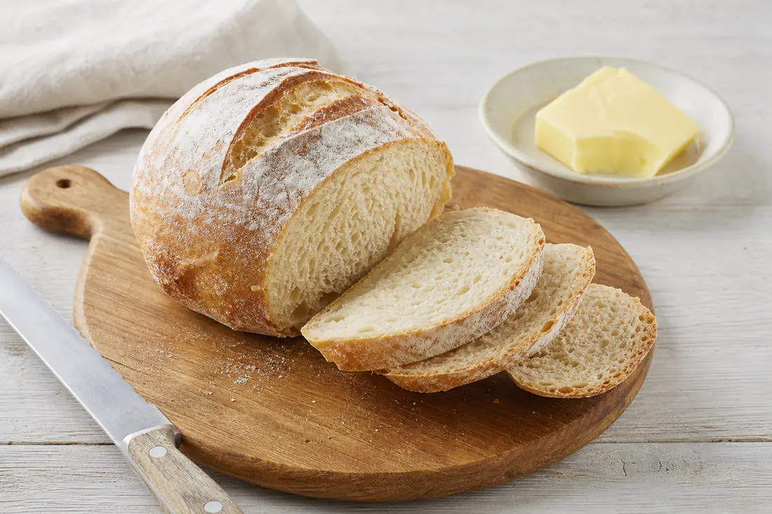 A small round white loaf with a golden crust and a floured top, sliced on a wooden board with butter