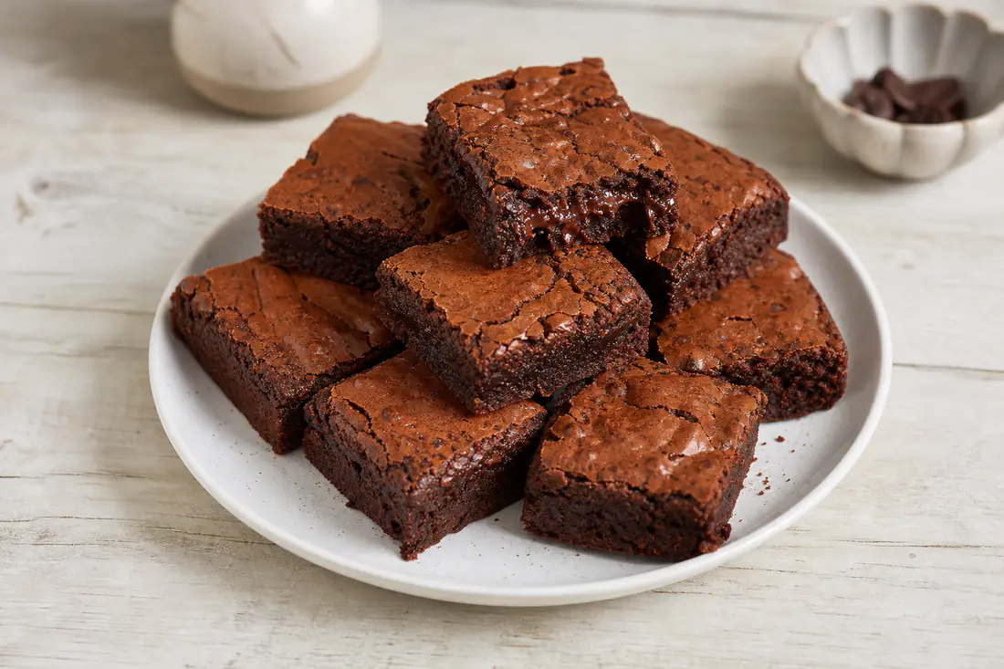 Nine square fudgy chocolate brownies with a crackly top stacked on a white plate