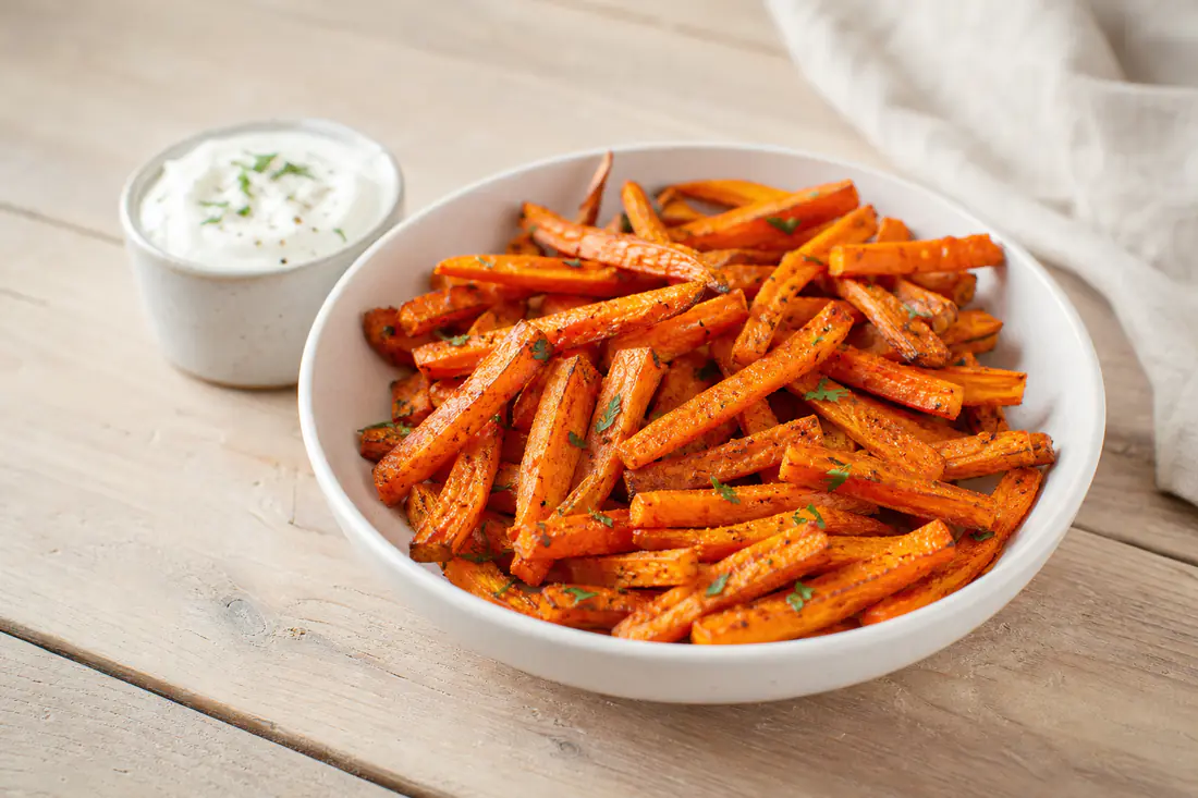 A white bowl of golden carrot fries sprinkled with paprika next to a small pot of garlic dip on a wooden table