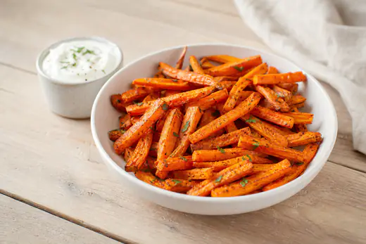 A white bowl of golden carrot fries sprinkled with paprika next to a small pot of garlic dip on a wooden table