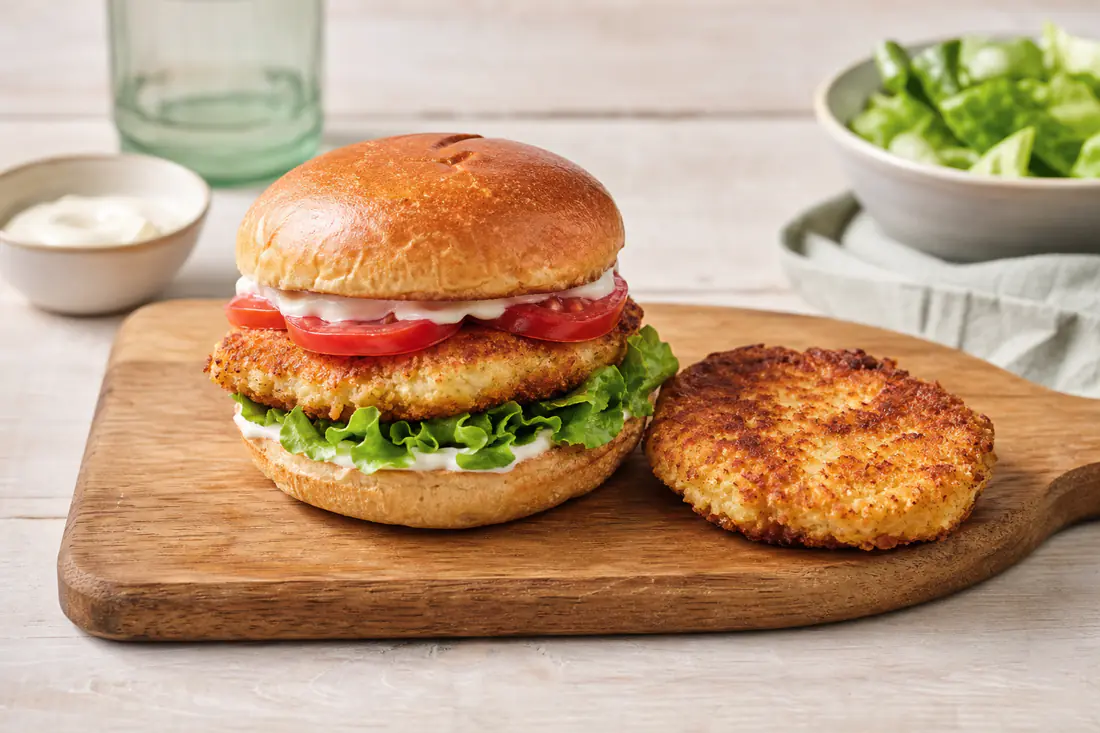 A chicken burger in a toasted bun with lettuce, tomato and mayonnaise on a wooden board next to a golden patty