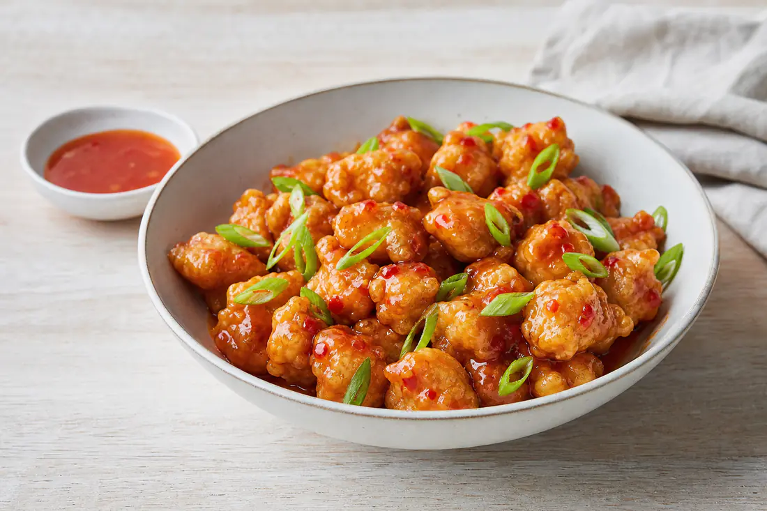 A bowl of golden crispy chicken bites glazed with sweet chilli sauce, with a small dish of dipping sauce and spring onion
