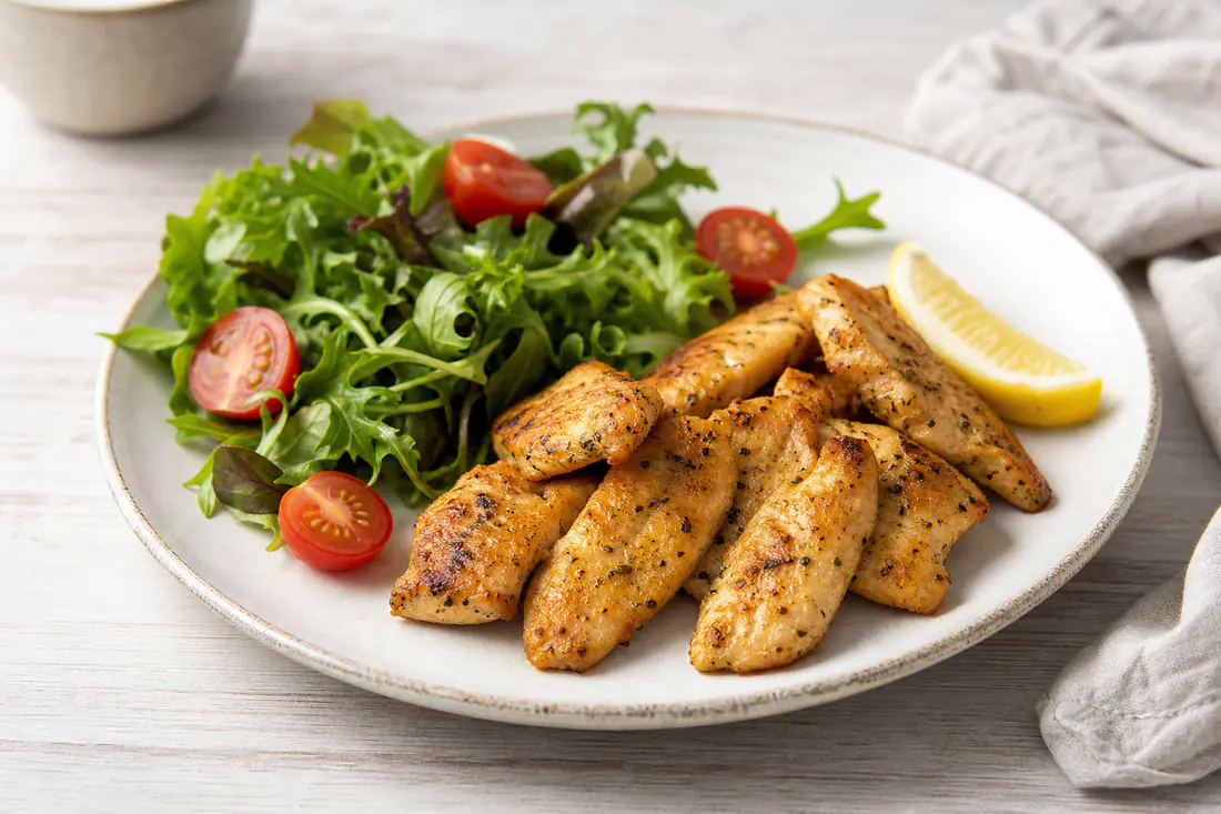 A plate of small golden seasoned chicken mini fillets with a lemon wedge, cherry tomatoes and a green salad