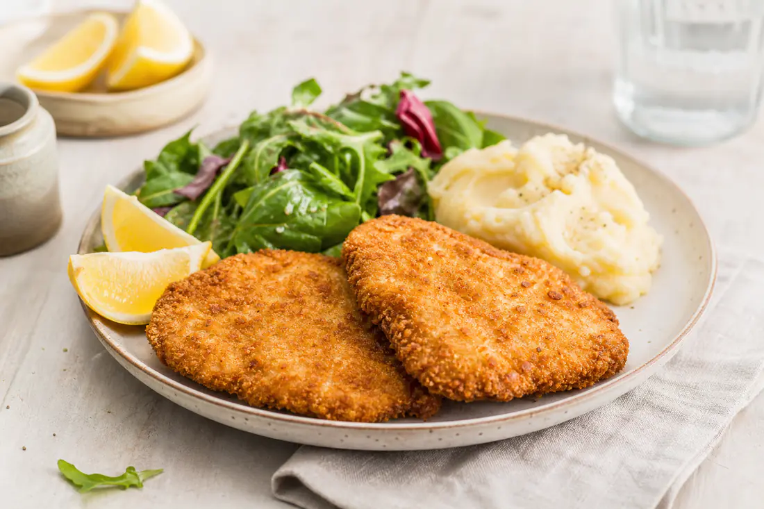Two golden crumbed chicken schnitzels on a plate with lemon wedges, a green salad and mashed potato