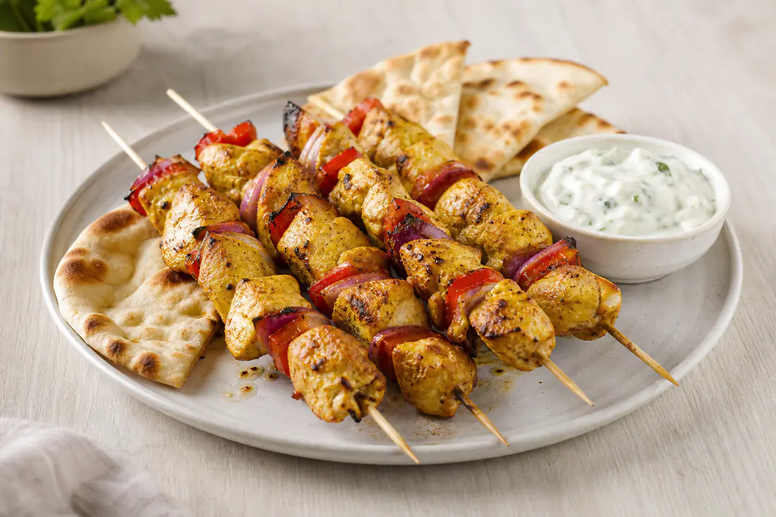 Four golden chicken skewers with red pepper and red onion on a plate with flatbread and a bowl of yoghurt dip