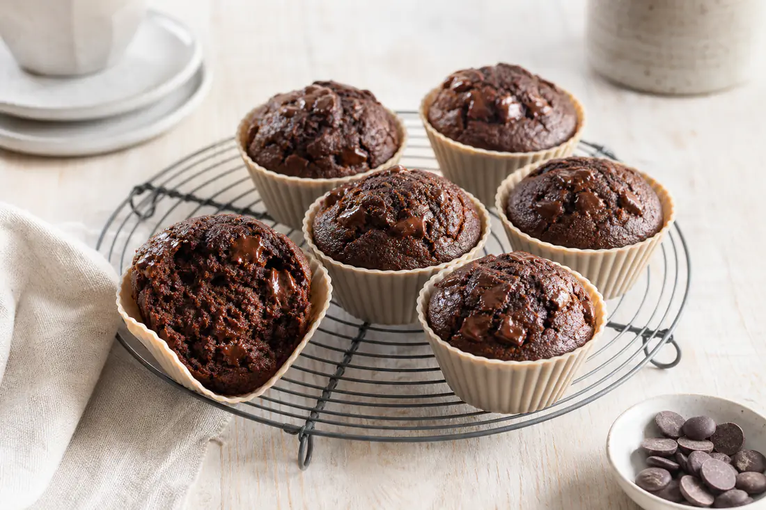 Six dark chocolate muffins with melted chocolate chips in silicone cases on a wire rack, one broken open to show the soft crumb