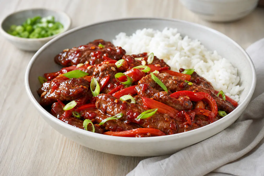 A bowl of crispy chilli beef strips glazed in red sauce with sliced peppers, spring onions and rice