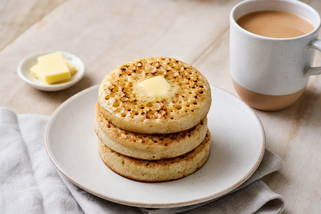 Three toasted crumpets with melted butter pooling in the holes, stacked on a white plate with a mug of tea