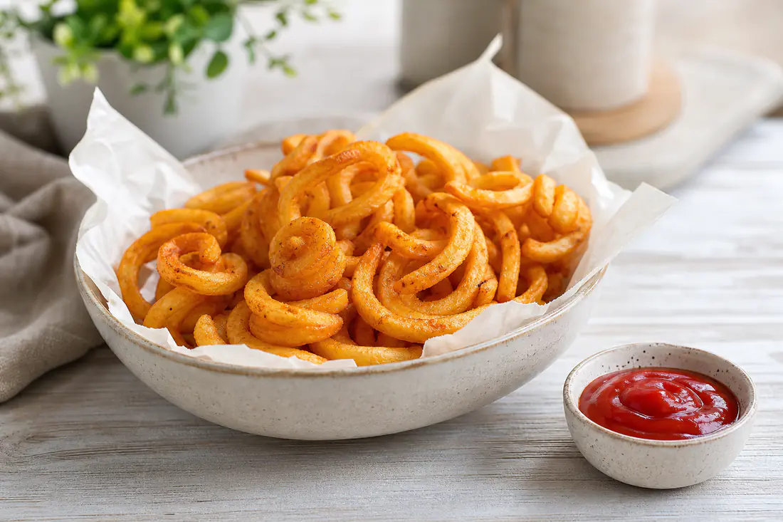 A paper-lined bowl of golden spiral curly fries with a small pot of ketchup on a kitchen table