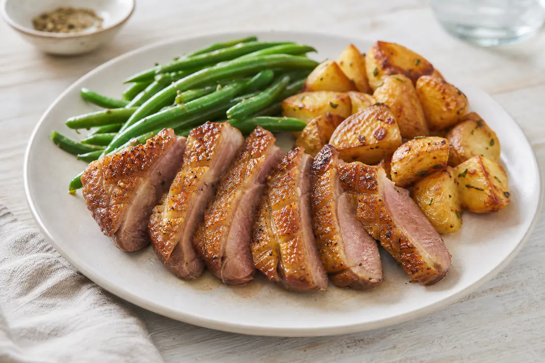 Sliced duck breast with crisp golden scored skin fanned out on a plate beside green beans and roast potatoes
