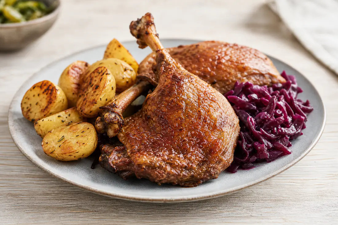 Two crisp golden roast duck legs with dark crackling skin on a plate with roast potatoes and red cabbage