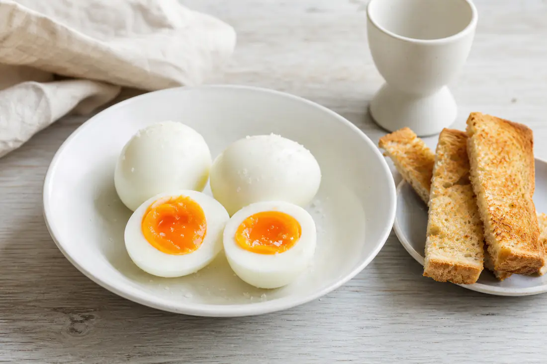 Four peeled boiled eggs, two halved to show soft yolks, in a white egg cup and bowl with toast soldiers