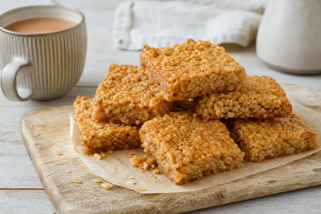 Golden oat flapjack squares stacked on a wooden board next to a mug of tea