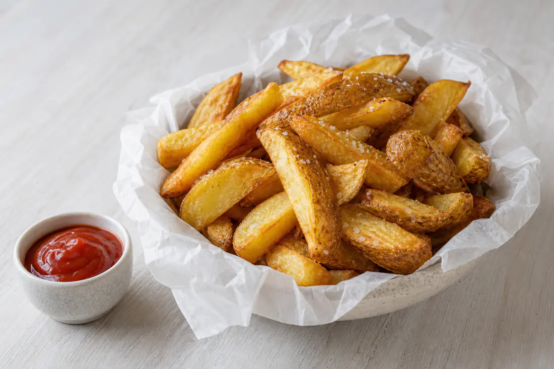 A paper-lined bowl of golden chunky homemade chips with sea salt, with a small pot of ketchup