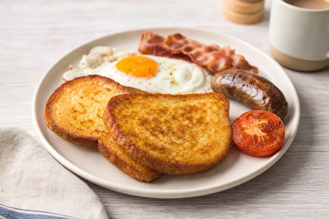 Two golden crisp slices of fried bread on a plate of full English breakfast with egg, bacon and grilled tomato