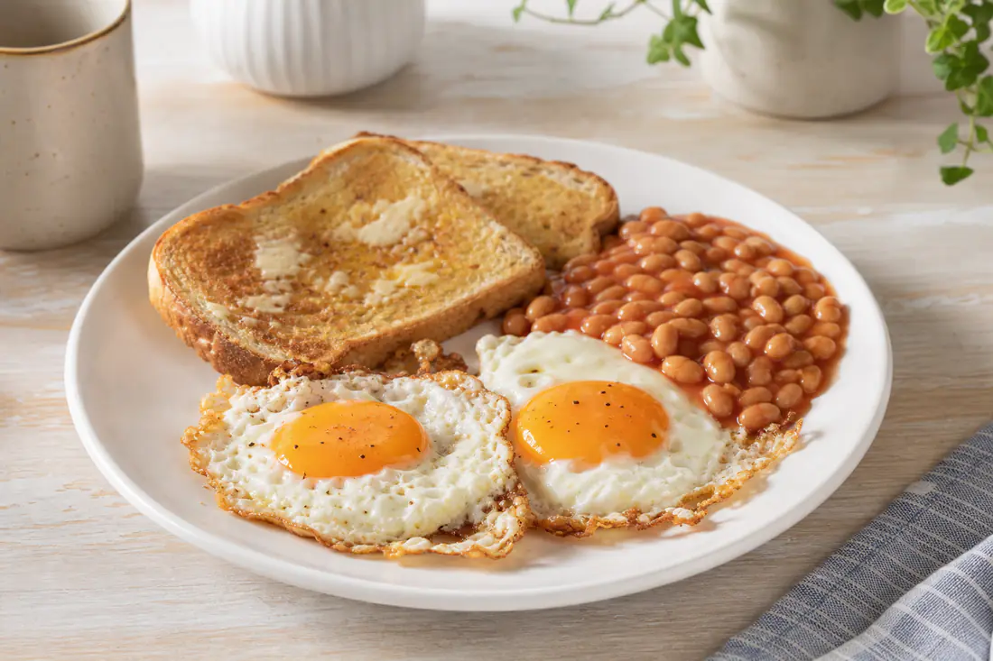Two sunny-side-up fried eggs with golden crisp edges and bright yolks on a plate with toast and baked beans