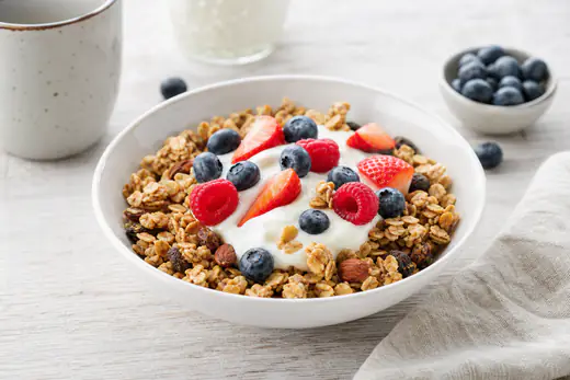 A white bowl of golden oat granola with nuts and raisins topped with yoghurt and fresh berries