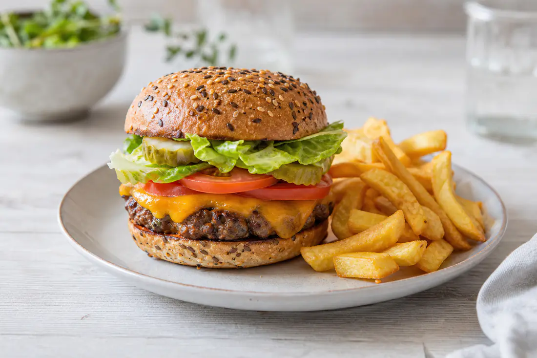 A beef burger in a seeded bun with melted cheddar, lettuce, tomato and sliced gherkin on a plate