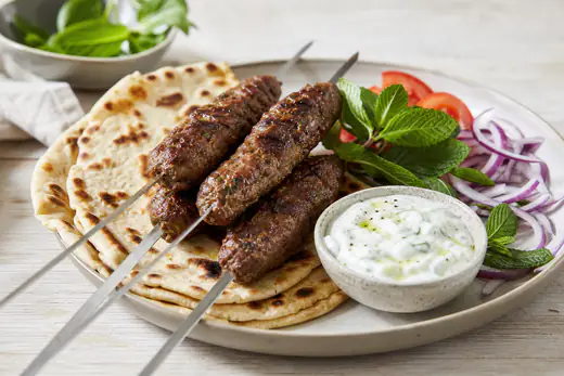 Browned lamb kofta kebabs on skewers on a plate with flatbread, yoghurt dip, sliced onion and tomato