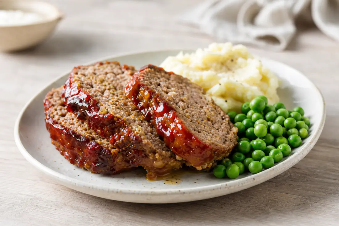 Thick slices of glazed meatloaf on a plate with mashed potato and peas