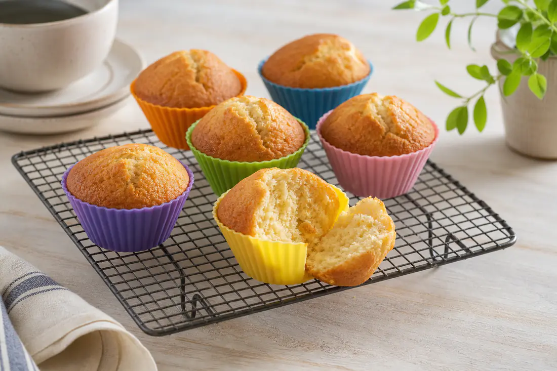 Six golden vanilla muffins in colourful silicone cases on a wire cooling rack, one broken open to show the soft crumb