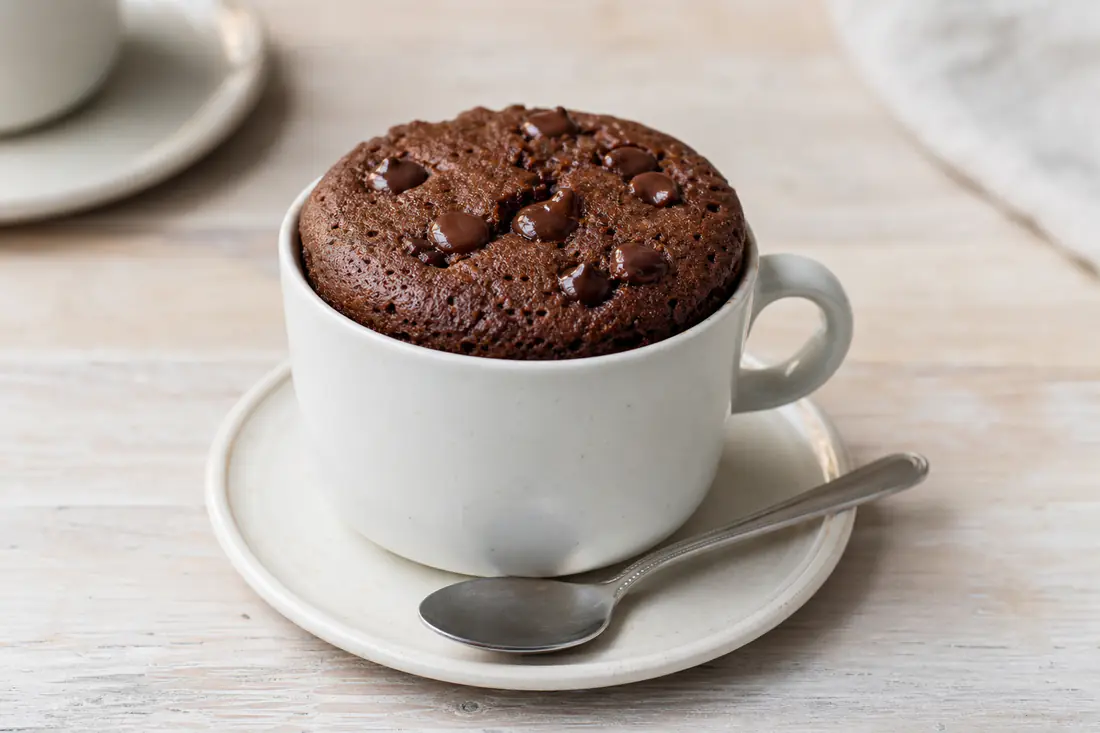 A chocolate mug cake risen in an ovenproof white mug with a spoon and chocolate chips on top
