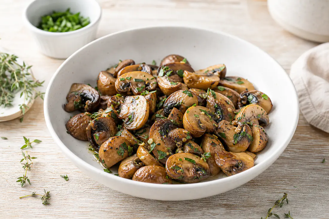 Golden roasted chestnut mushrooms with garlic and thyme in a white serving bowl on a wooden table