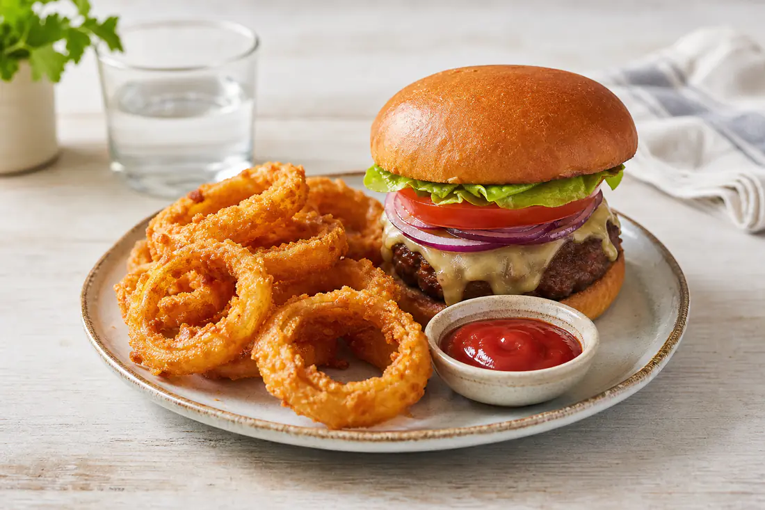 A plate of golden crispy breaded onion rings stacked beside a small bowl of ketchup and a burger on a kitchen table