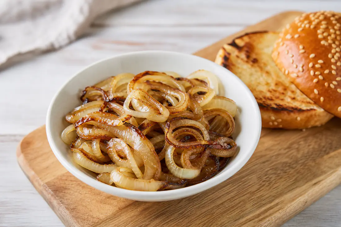 Golden fried onion slices piled in a white bowl beside a burger bun on a wooden board