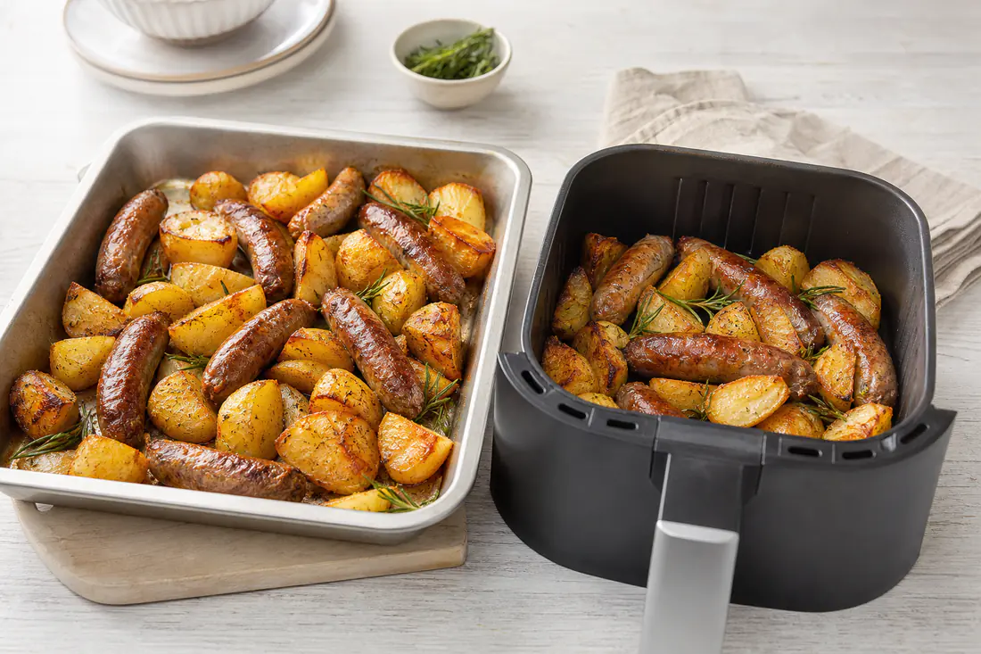 Golden roast potatoes and sausages in an air fryer basket beside a baking tray of the same food from the oven