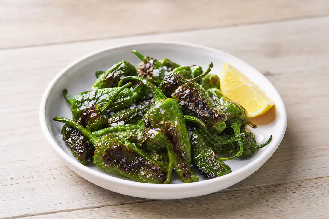 Blistered green padron peppers with flaky sea salt in a small white dish with a lemon wedge on a wooden table