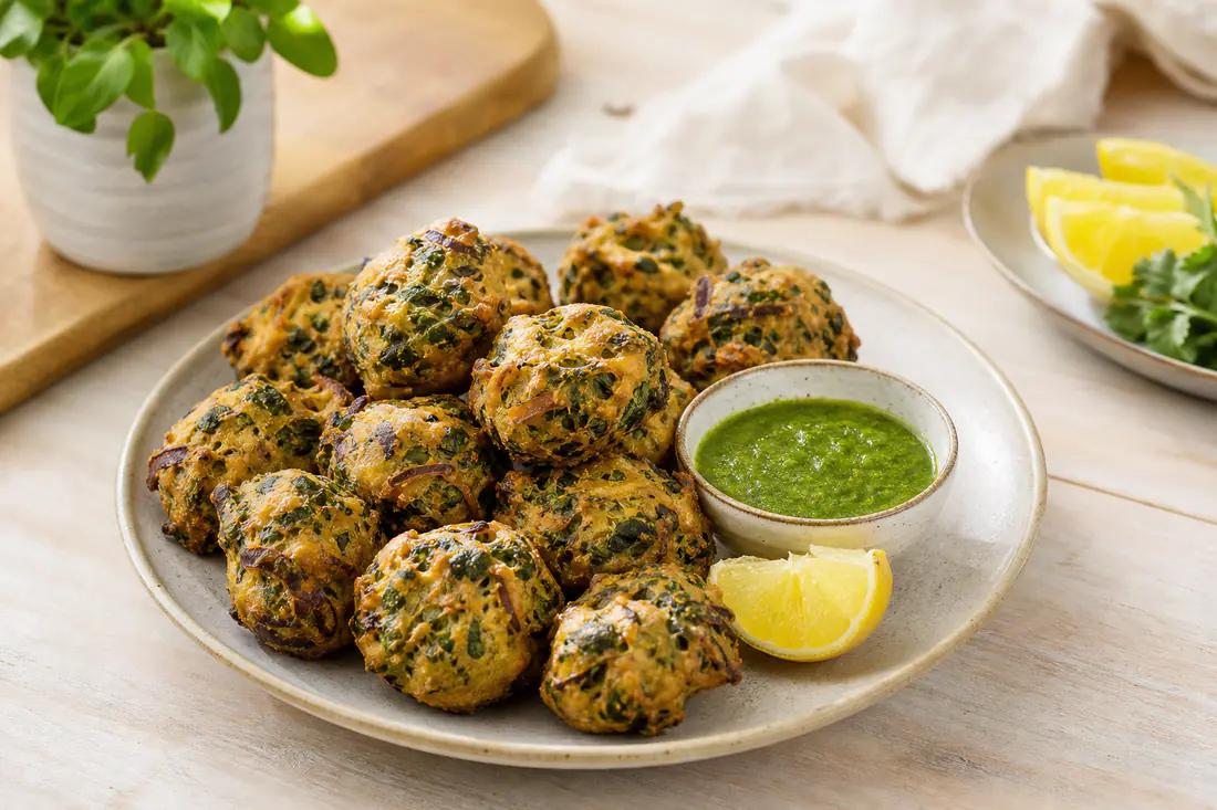 Golden vegetable pakoras with spinach and potato on a serving plate with mint chutney and lemon wedges