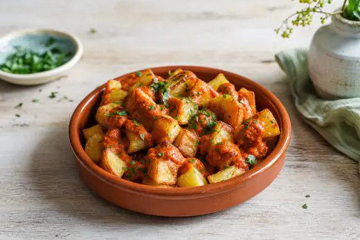 A terracotta tapas dish of golden crispy potato cubes topped with red smoky bravas sauce and chopped parsley