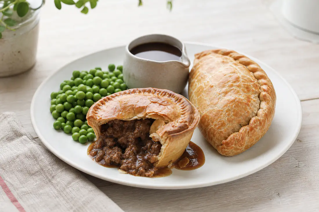 A golden individual steak pie cut open to show the meat filling next to a Cornish pasty on a plate with peas