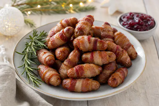 Golden pigs in blankets, chipolatas wrapped in crisp streaky bacon, on a white plate with a sprig of rosemary