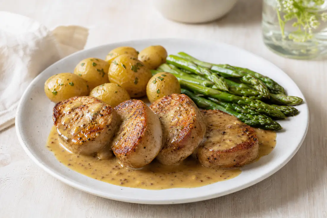 Browned pork medallions on a plate with a mustard sauce, new potatoes and asparagus