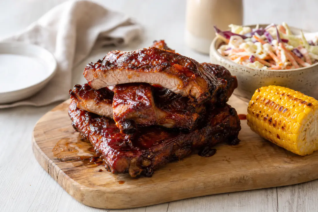A stack of glazed pork spare ribs with sticky barbecue sauce on a wooden board with coleslaw and corn