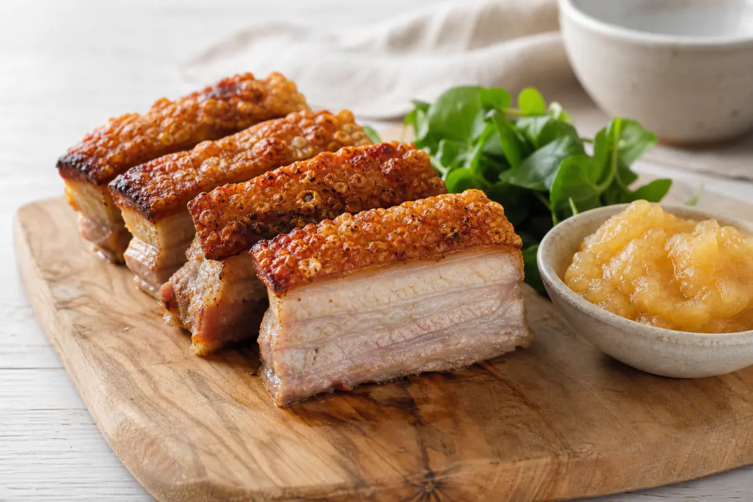 Thick slices of pork belly with golden blistered crackling on a wooden board with apple sauce and watercress