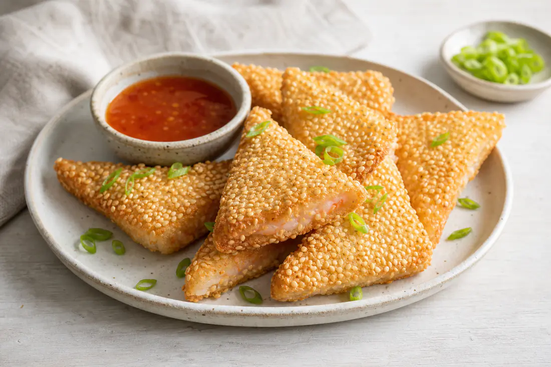 Golden sesame prawn toast triangles on a plate with sweet chilli sauce and sliced spring onion