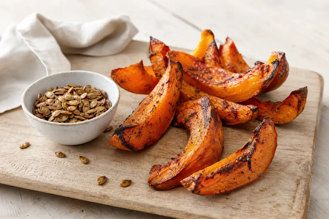 Roasted orange pumpkin wedges with dark edges and a small bowl of roasted pumpkin seeds on a board